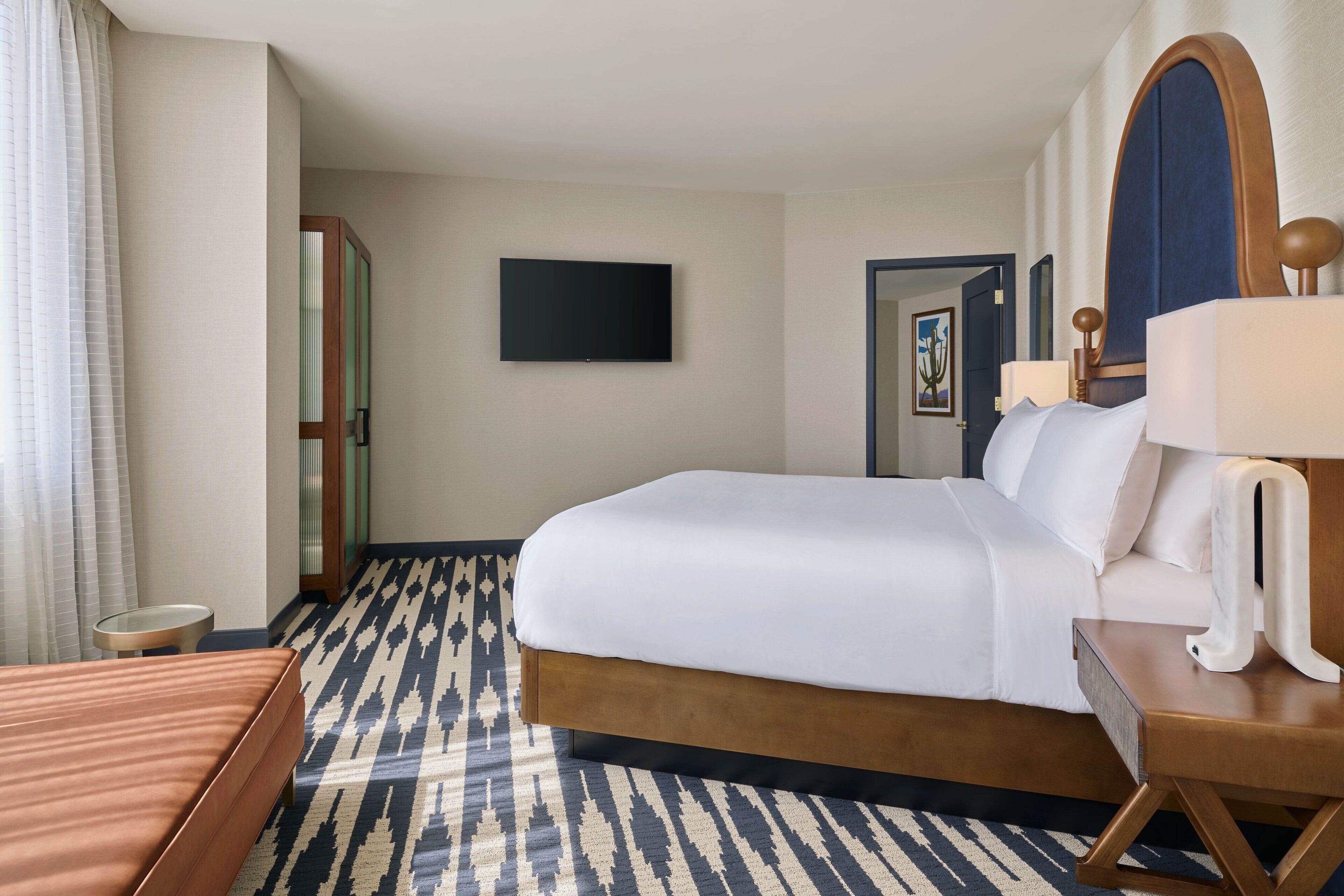 A hotel room with a large bed, a bedside table with a lamp, a flat-screen TV mounted on the wall, and a patterned carpet.