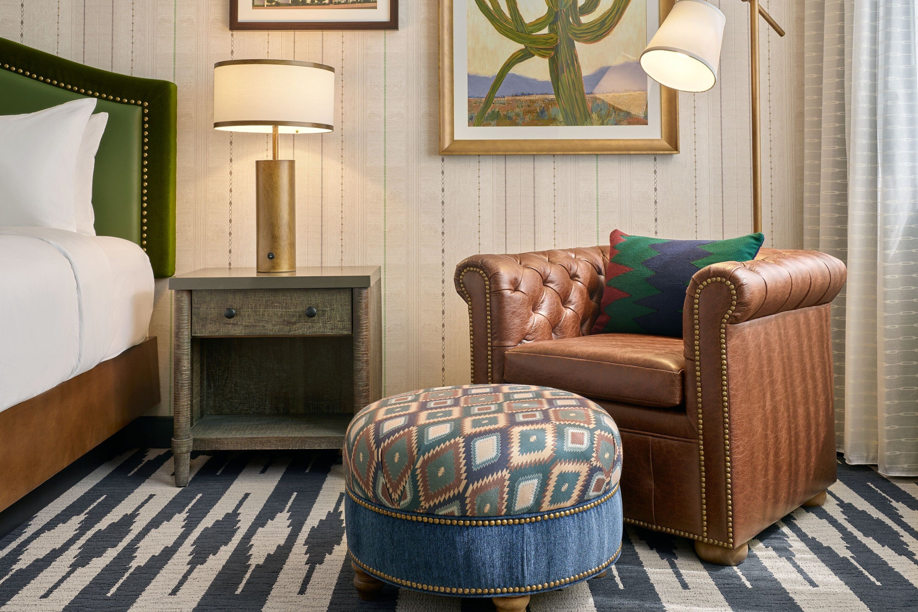 A hotel room with a bed, a nightstand, a leather armchair, a patterned ottoman, a lamp, and a framed picture on the wall.