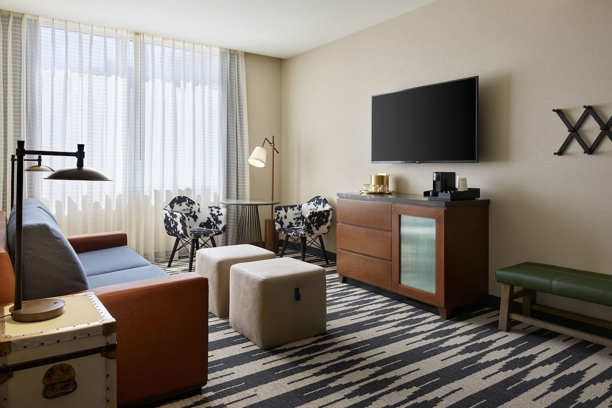 A modern hotel room with a sofa, ottoman, a small table, a TV mounted on the wall, and a cabinet.