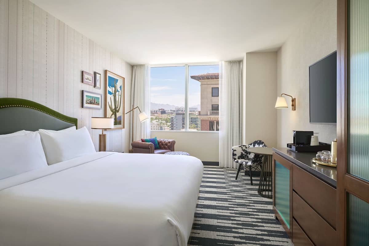 A hotel room with a large bed, a desk with a chair, a TV, and a window with a city view.