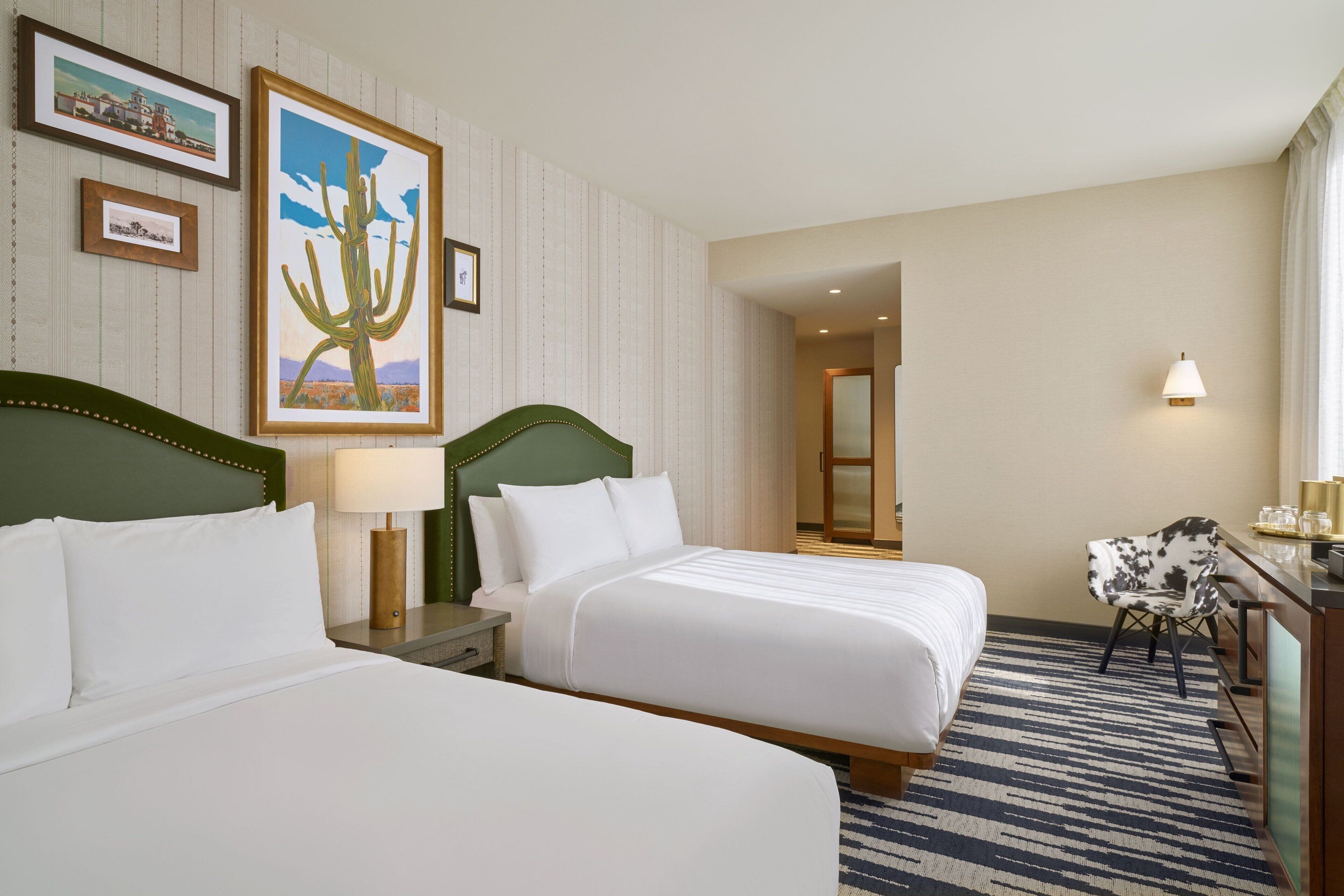 A hotel room with two beds, a desk, a chair, and a wall with framed pictures.