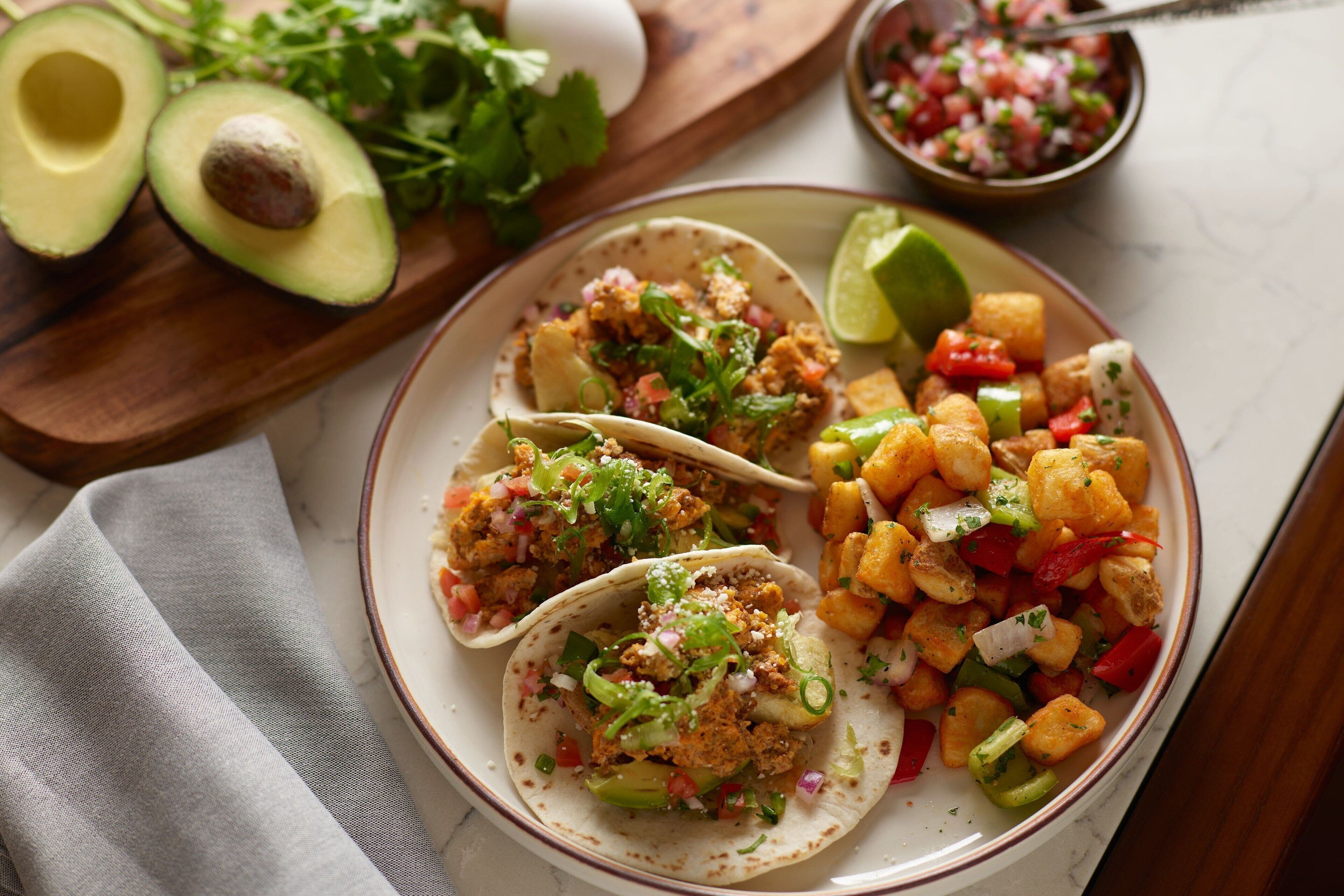 Een bord met drie taco's, een portie blokjes groenten en halve avocado's op een houten plank.