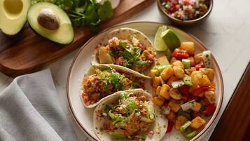Un plato con tres tacos, una guarnición de verduras picadas y medio aguacate sobre una tabla de madera.