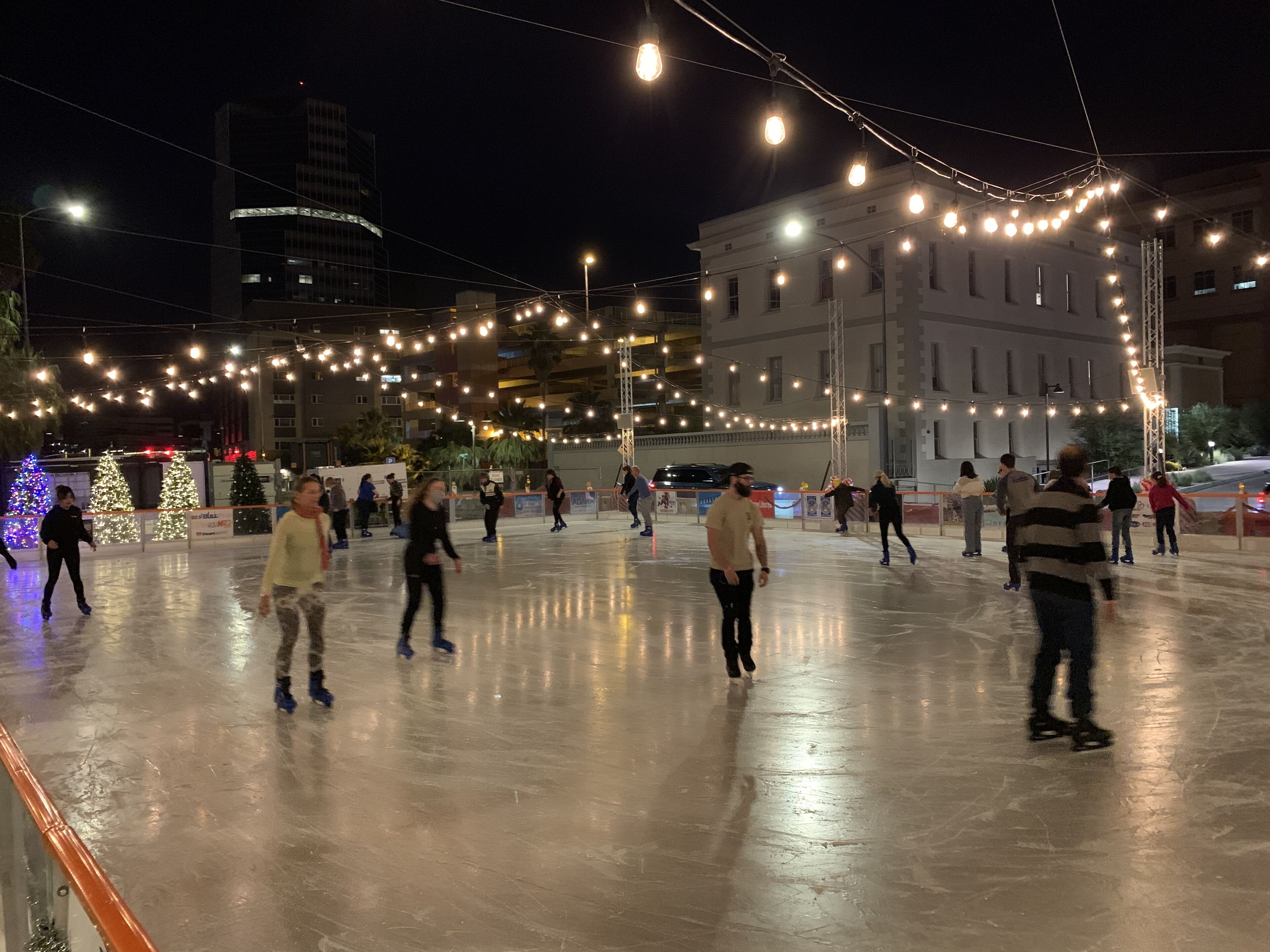 An outdoor ice skating rink with people skating, surrounded by buildings and string lights.