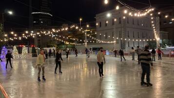 An outdoor ice skating rink with people skating, surrounded by buildings and string lights.