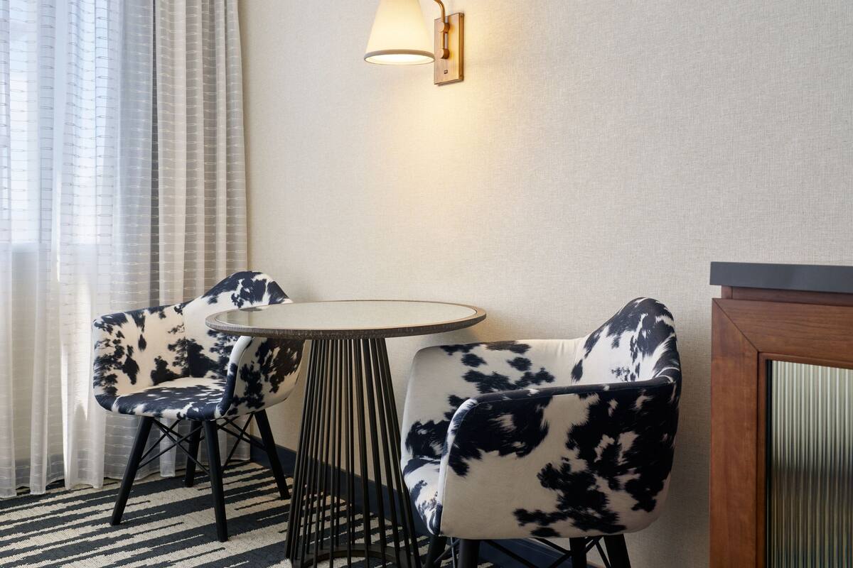 A room with a round table and two chairs with a black and white pattern, a wall-mounted lamp, and a window with sheer curtains.
