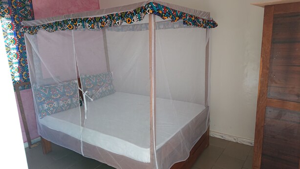 Un letto con zanzariera e cuscini decorati, una porta in legno e un pavimento piastrellato.