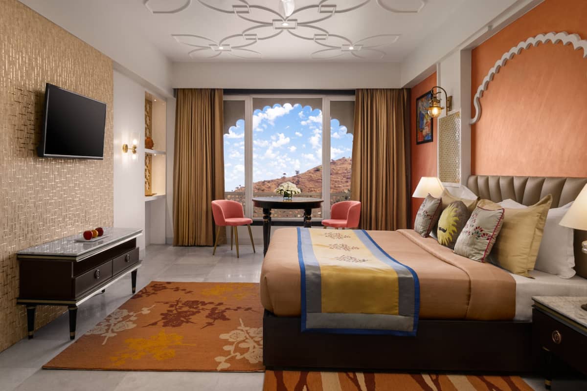 A hotel room with a bed, a desk, a chair, a TV, and a view of the mountains.