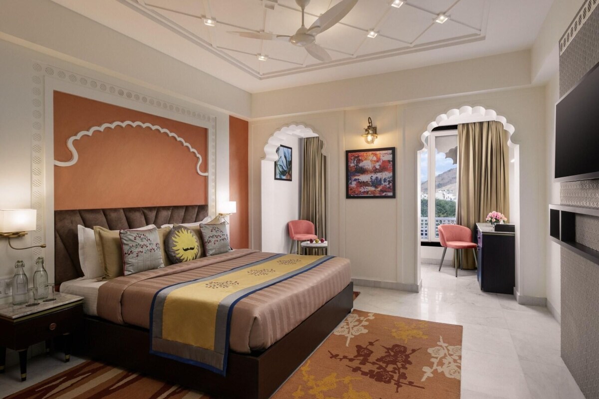 A hotel room with a large bed, a desk, a chair, a television, and a balcony with curtains.