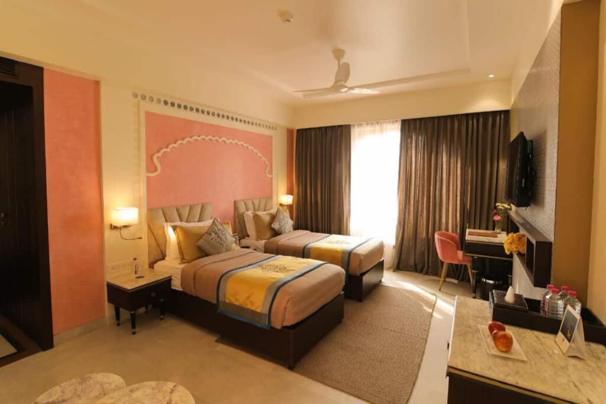 A hotel room with two beds, a desk, a chair, a TV, and a ceiling fan.