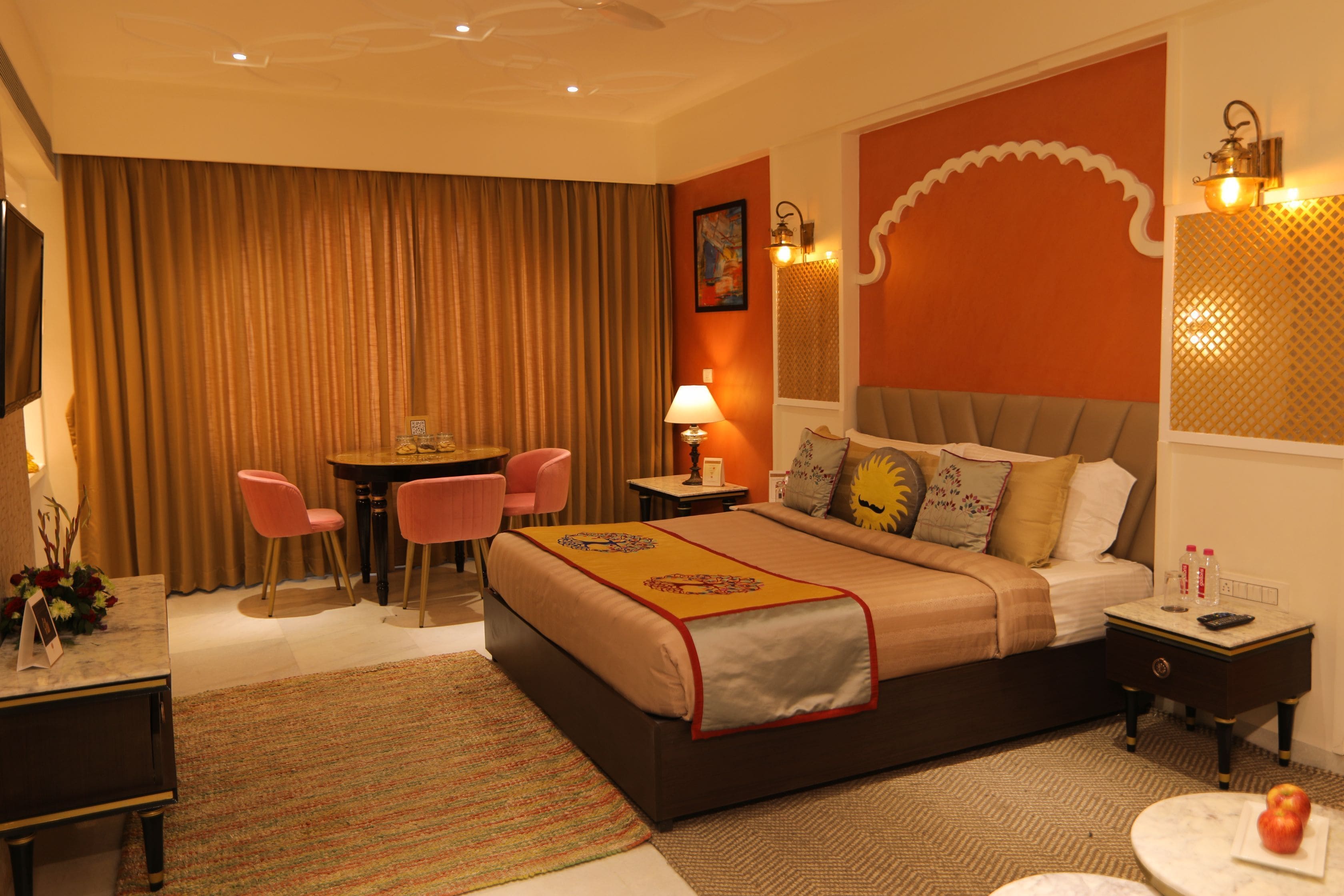 A hotel room with a large bed, a desk with a chair, a small table, and a nightstand with a lamp.