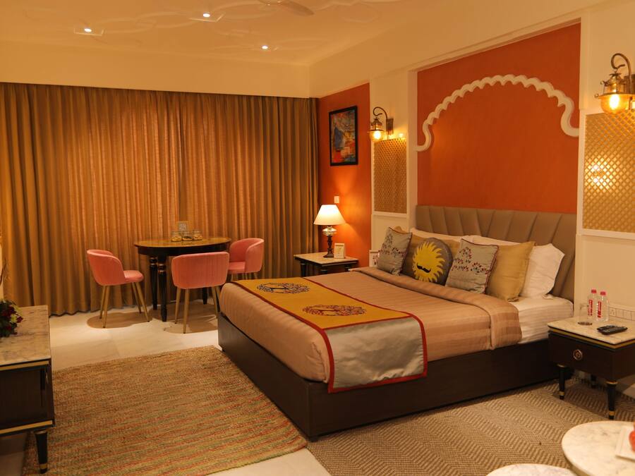 A hotel room with a large bed, a desk with a chair, a small table, and a nightstand with a lamp.