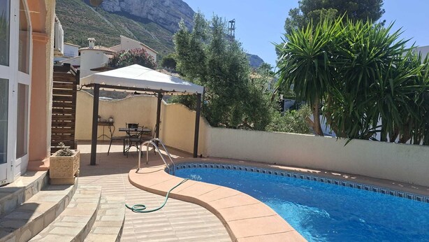 A swimming pool with a curved edge, a covered patio with outdoor furniture, and a mountainous backdrop. - Cosy villa with private pool and sea and mountain views, in Denia (Denia)