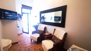 A room with a wooden floor, a brown leather chair, a white armchair, a small table with a vase, and a wall-mounted TV displaying various streaming services. - Frutas do Mar apartamento (Portimão)