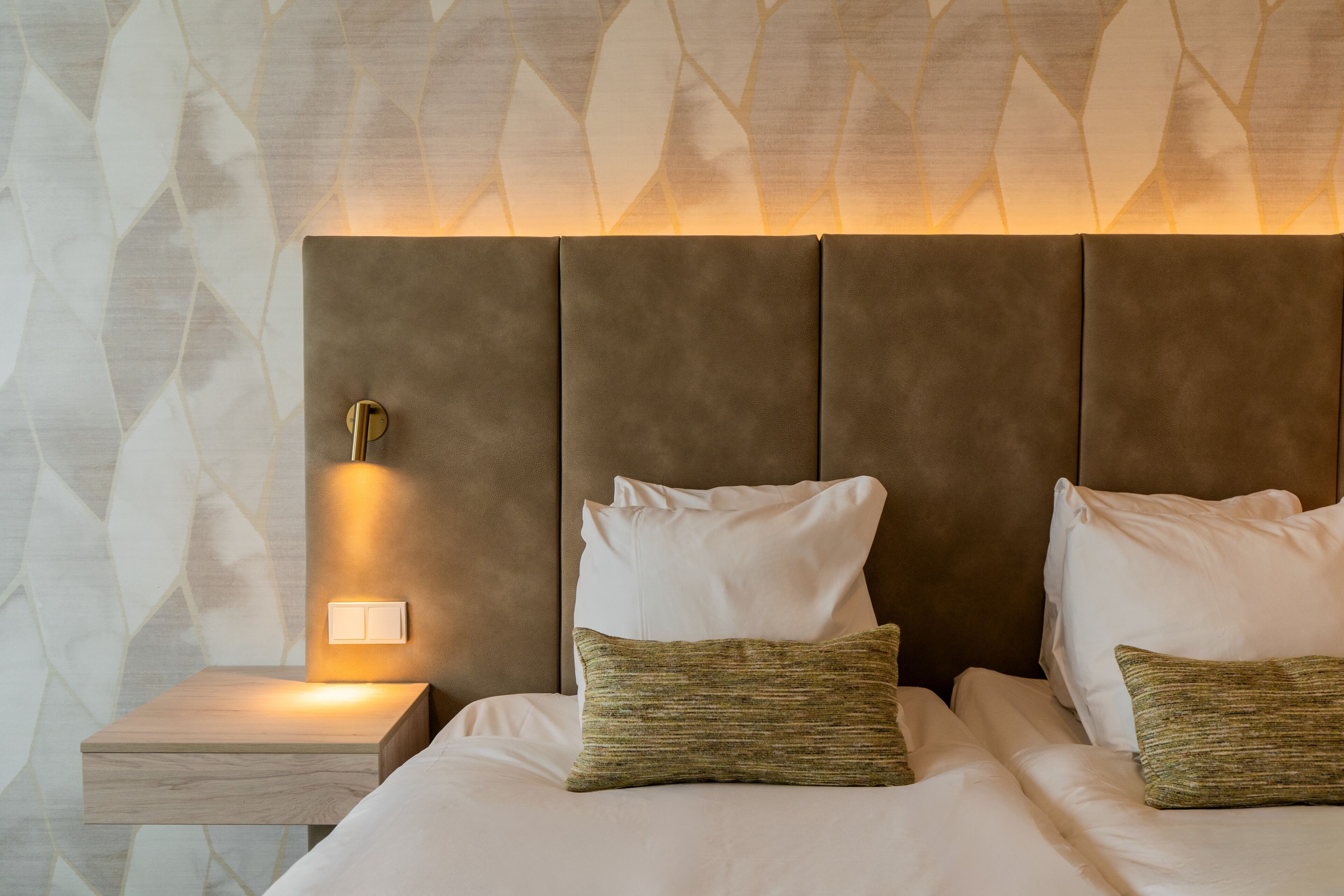 Un lit bien fait, avec des oreillers blancs et verts, une lampe de chevet et un mur orné d’un motif de feuilles.