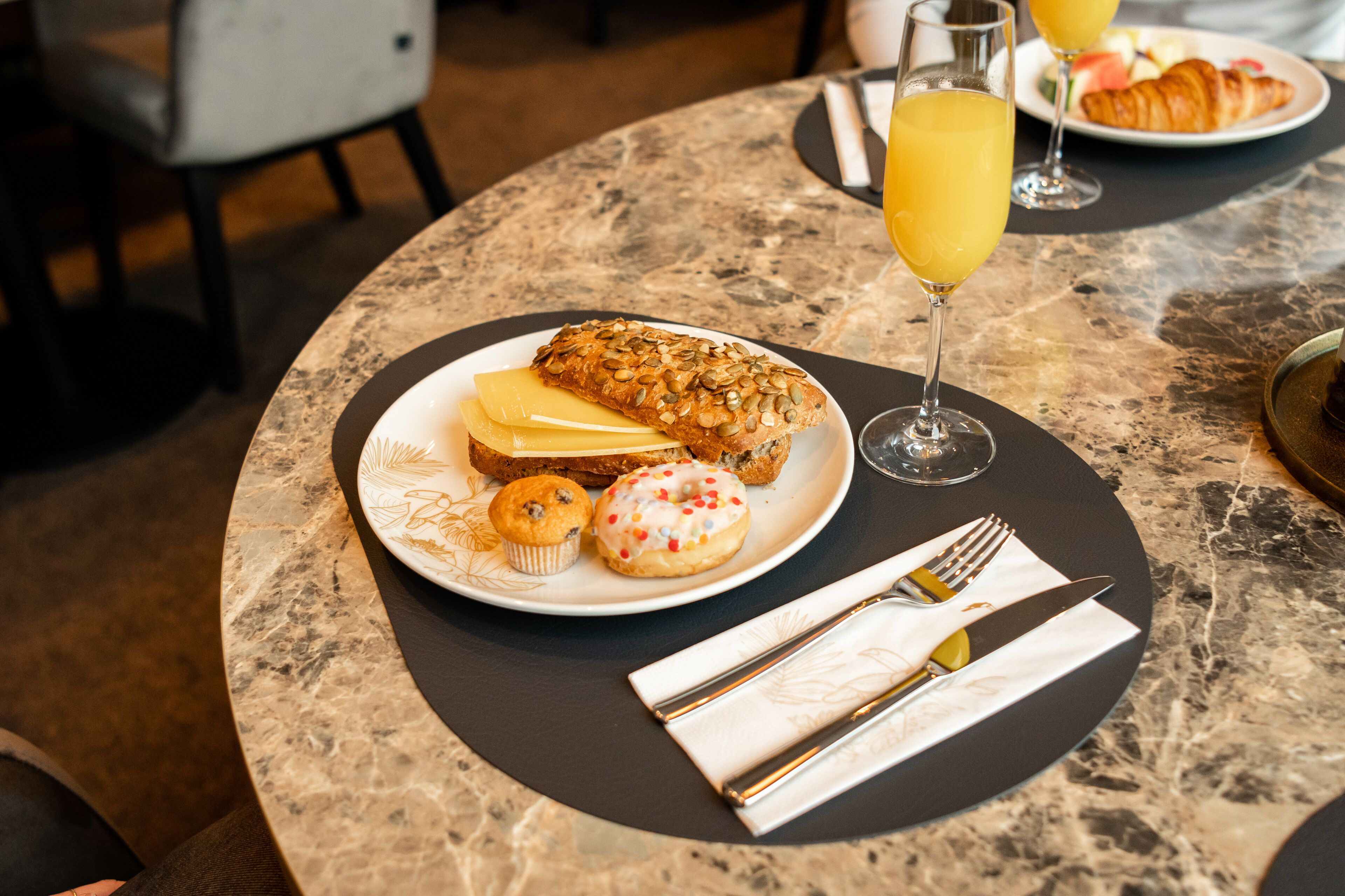 A breakfast setting with a croissant, muffin, and donut on a plate, a glass of orange juice, and cutlery on a marble table.