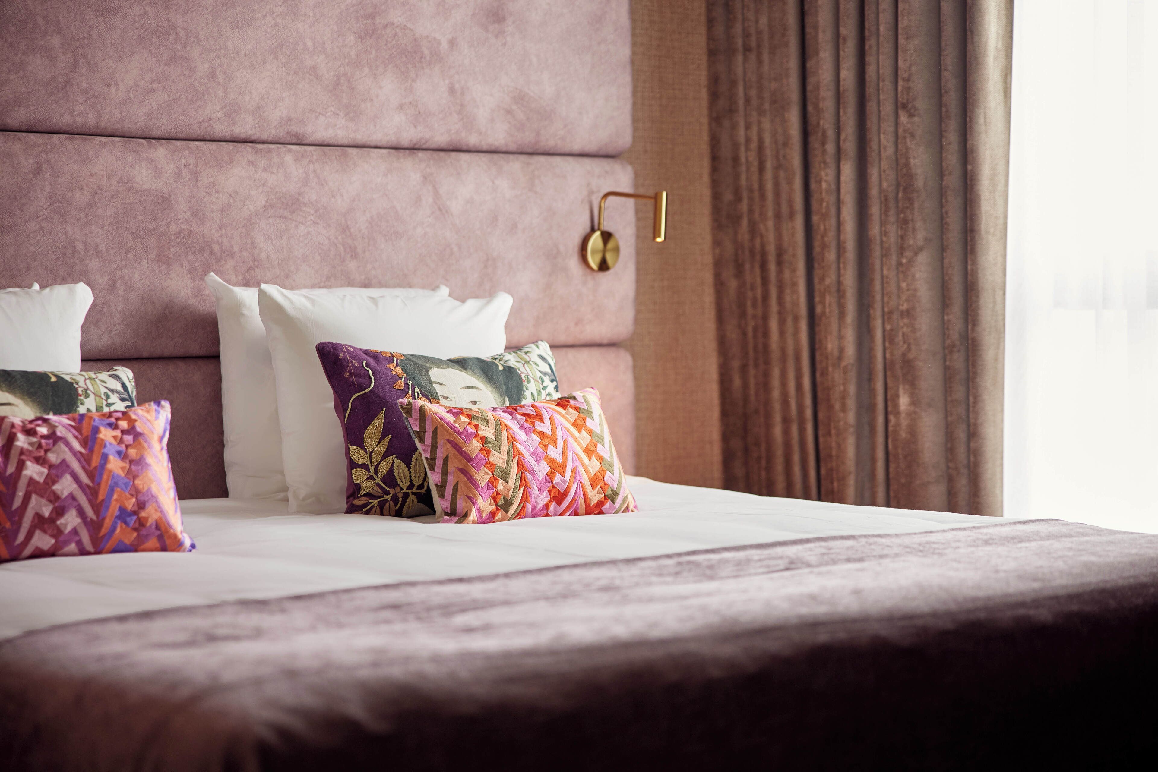 Una cama bien hecha con un cabecero rosa, dos almohadas blancas y almohadas estampadas de colores.