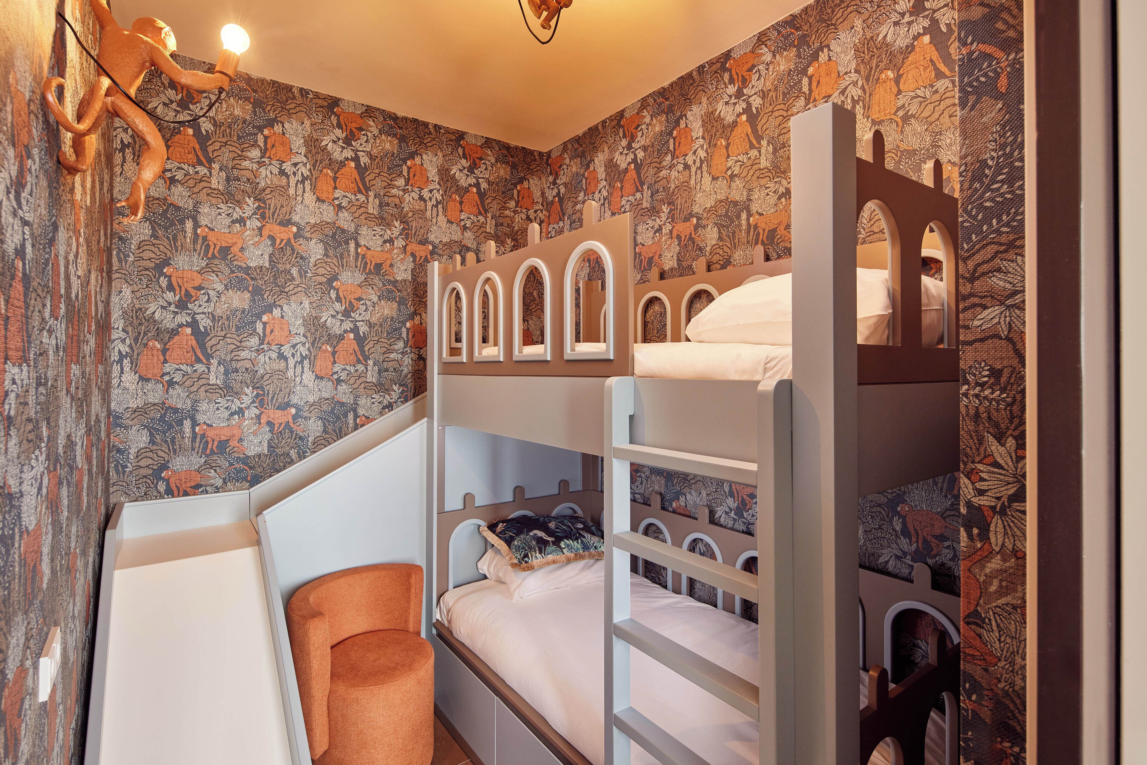 A bunk bed room with a floral wallpaper, a small orange chair, and a wooden shelf.