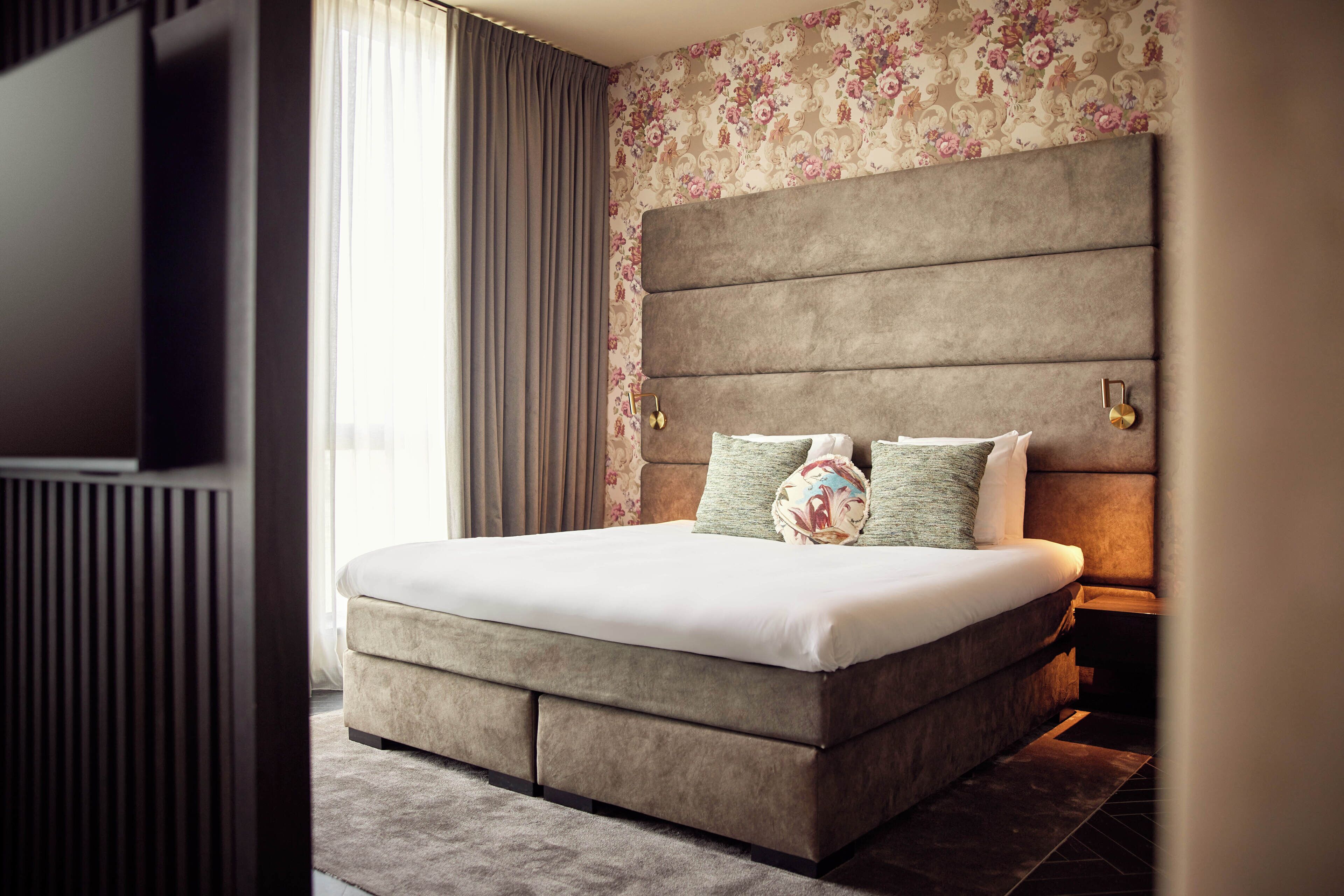 A modern bedroom with a large bed, a headboard with a built-in TV, and a floral wallpaper.