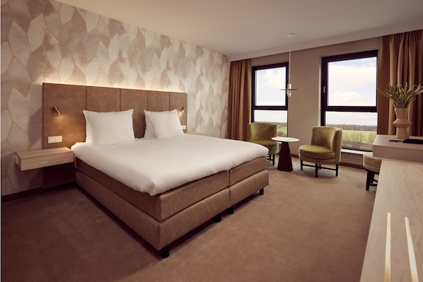 A modern hotel room with a large bed, a nightstand, a small round table, two armchairs, and a view of the countryside through large windows. - Van der Valk Hotel Gorinchem - A15 (Gorinchem)