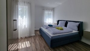 A bedroom with a bed, two windows with curtains, a bedside table, and a wall-mounted light fixture. - "achterwasser" - Moderne Ferienwohnung in Varel (Varel)