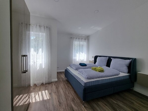A bedroom with a bed, two windows with curtains, a bedside table, and a wall-mounted light fixture. - "achterwasser" - Moderne Ferienwohnung in Varel (Varel)
