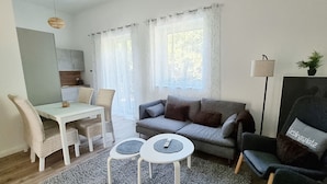 A living room with a sofa, two small tables, a dining table, and chairs. There is a lamp, a potted plant, and a window with curtains. - "steuerbord" - Moderne Ferienwohnung in Varel (Varel)