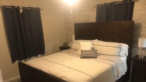 Una cama bien hecha con cabecera, una mesita de noche con lámpara y cortinas oscuras.