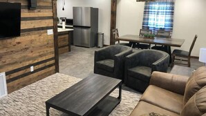 A modern living room with a sofa, armchairs, and a coffee table. There is a dining area with a table and chairs, and a kitchen with a refrigerator and microwave.