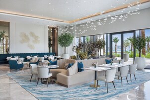 A modern hotel lobby with a seating area, a bar, and large windows offering a view of palm trees and a cityscape.