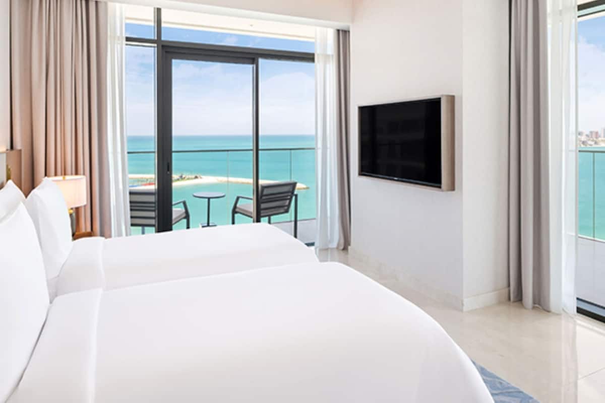 A hotel room with a large bed, a TV mounted on the wall, a balcony with a view of the sea, and a small table with chairs.