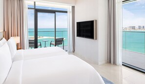 A hotel room with a large bed, a TV mounted on the wall, a balcony with a view of the sea, and a small table with chairs.