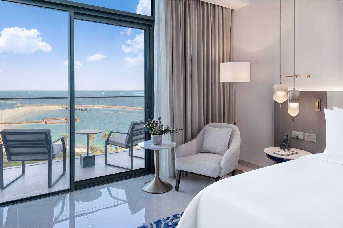 A hotel room with a large balcony, a bed, a chair, a small table with a vase, and a nightstand with a lamp and phone.