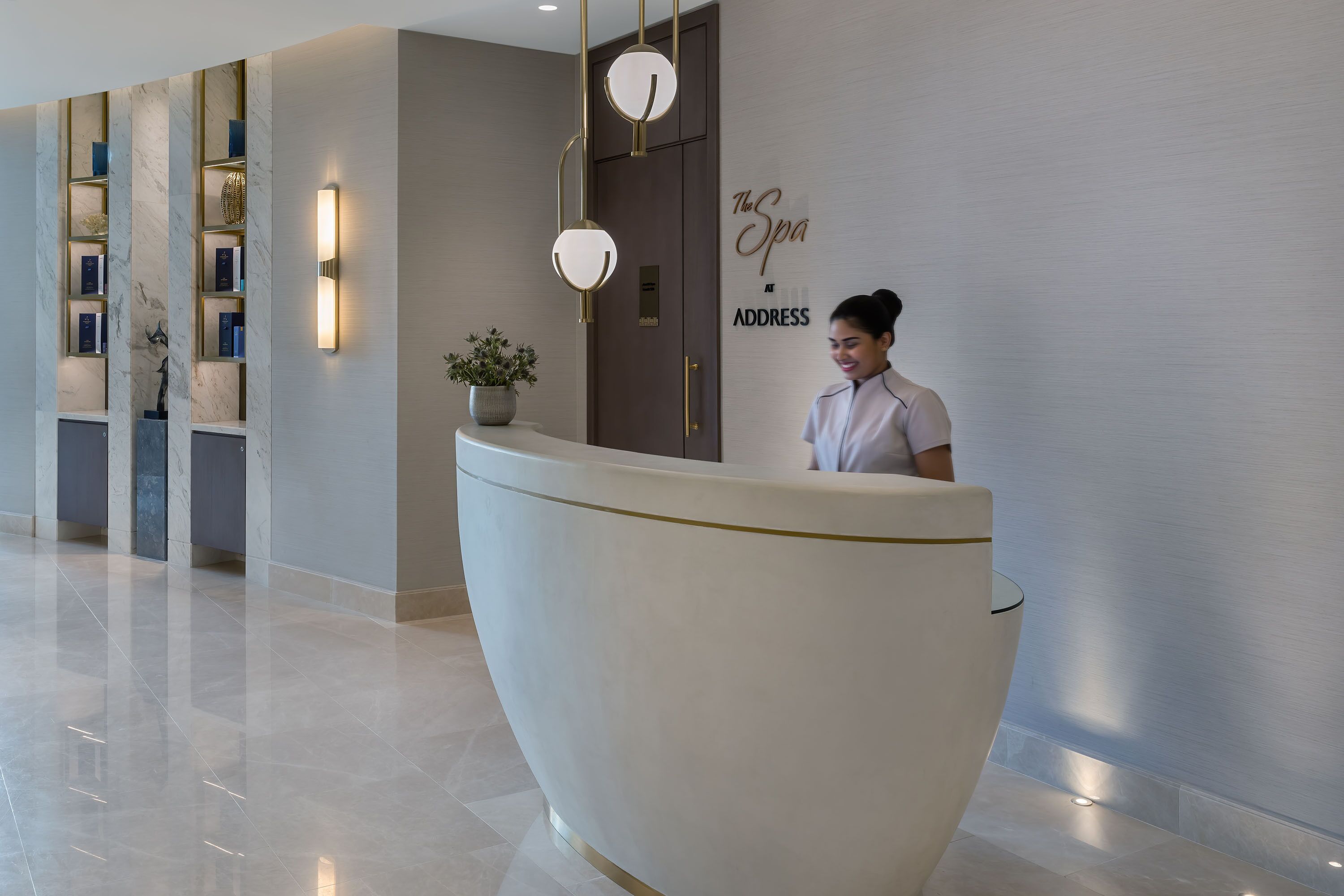 A reception desk with a modern design, a wall-mounted sign reading "The Spa at ADDRESS", and a reception area with a polished floor and decorative shelves.