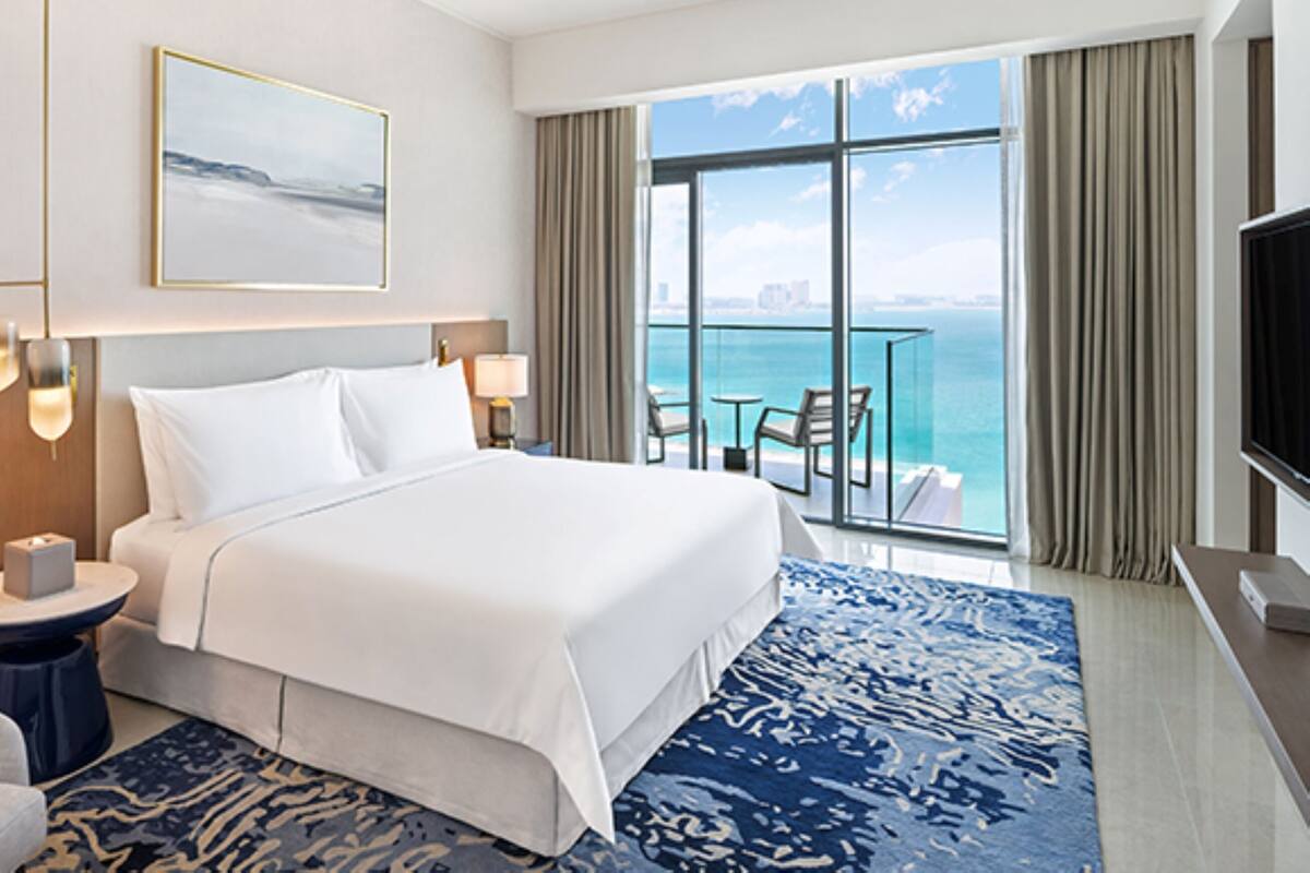 A hotel room with a large bed, a view of the ocean, a balcony with seating, and a television.