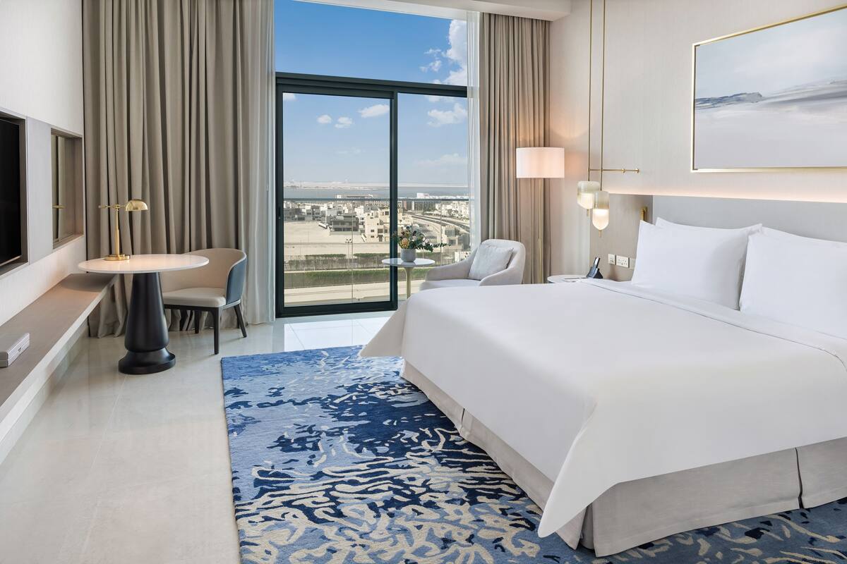 A hotel room with a large bed, a desk with a chair, a TV, and a view of the city.