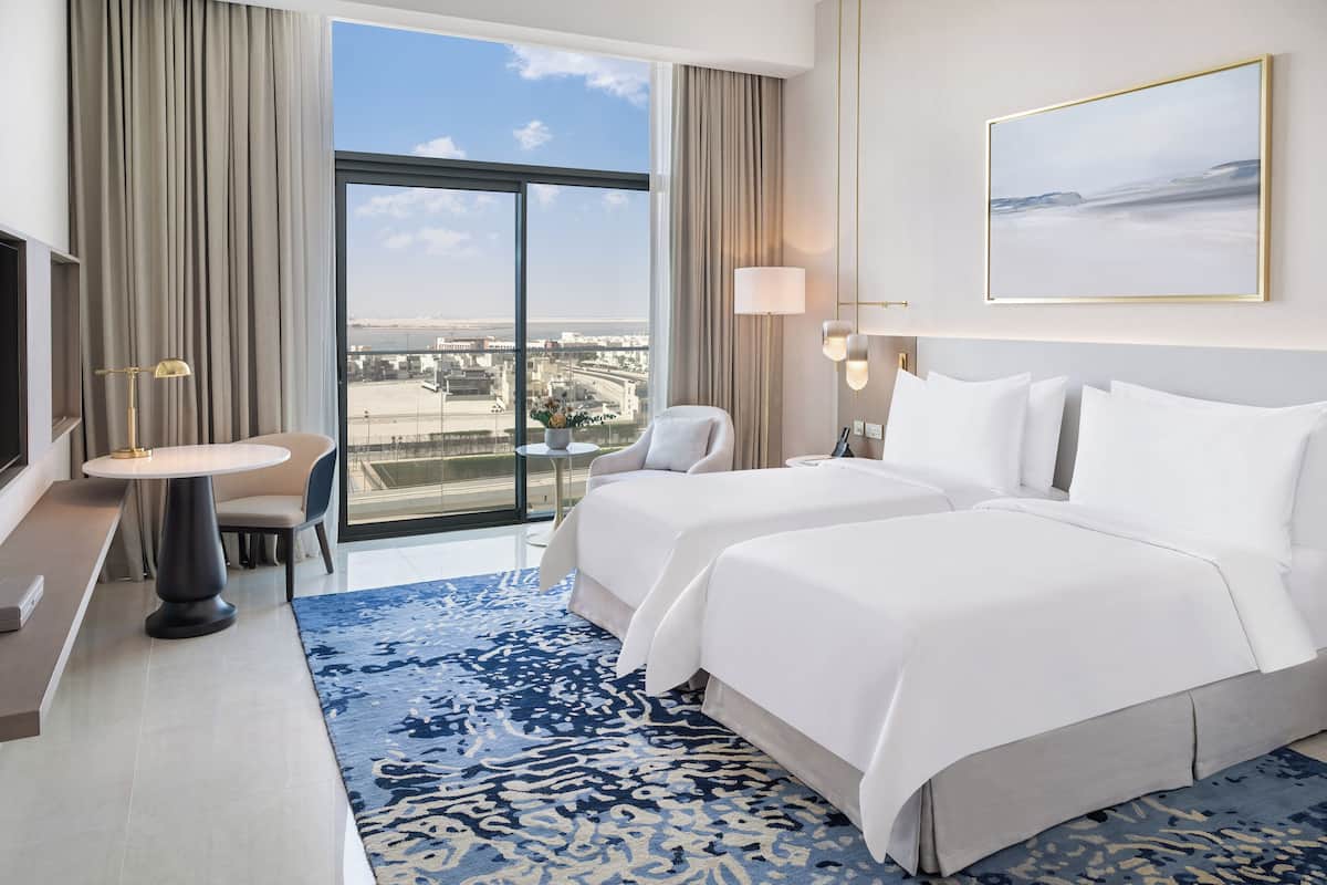 A hotel room with a large window, two beds, a desk, and a chair.