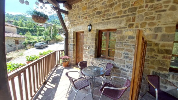 A stone building with a balcony, outdoor seating, and a view of a road and greenery.