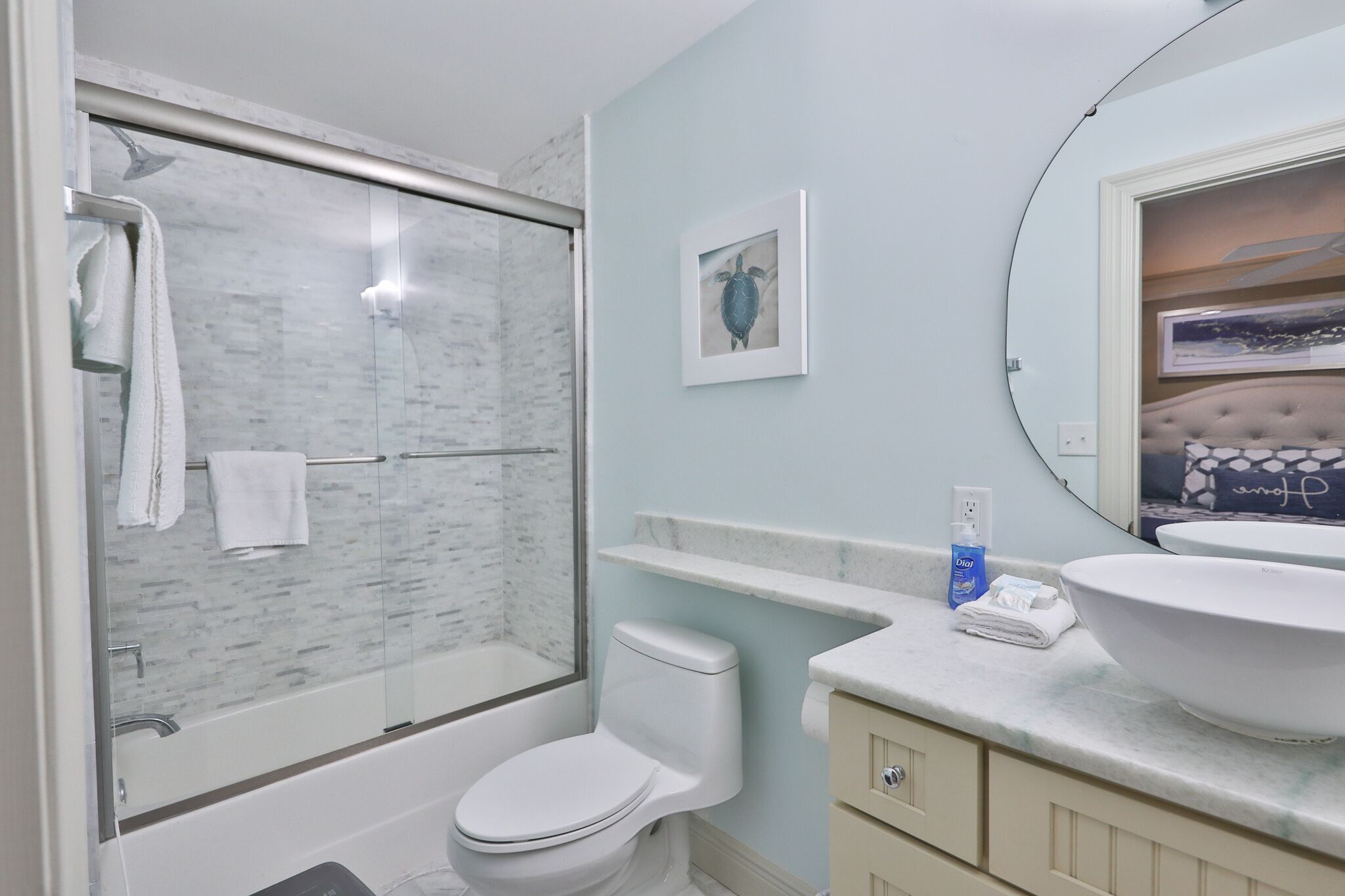 A bathroom with a glass shower enclosure, a white toilet, a large sink with a bowl, a vanity with a mirror, and a framed picture on the wall.