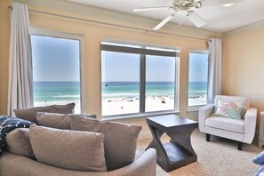 A living room with a view of the beach, a ceiling fan, and a chair with a cushion.