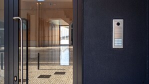 Entrada de um edifício moderno com porta de vidro, puxador metálico e um teclado de segurança na parede.