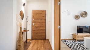 Um corredor com porta de madeira, um vaso alto com plumas de capim-do-campo, uma pequena mesa com uma planta, um abajur de chão preto e uma televisão sobre um suporte de madeira.