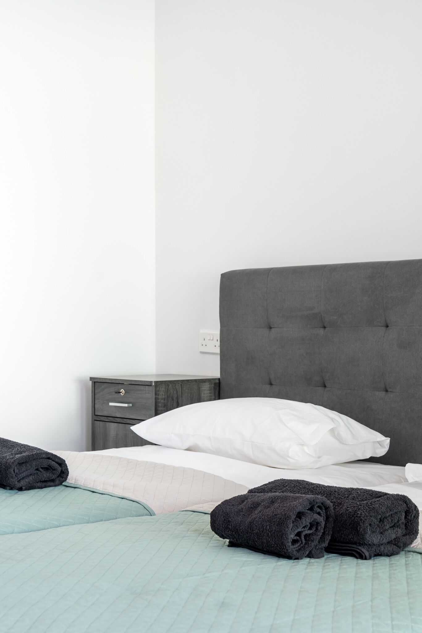 A neatly made bed with a grey headboard, a bedside table with a drawer, and a light green bedspread.