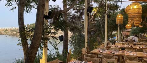 Un área de comedor al aire libre con mesas y sillas de madera, rodeada de árboles y con vista a un cuerpo de agua.