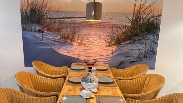 Een eettafel gedekt voor zes personen, met rieten stoelen, een houten tafelvoet en een schilderij van een zonsondergang boven het water op de achtergrond.