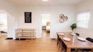Un espacio habitable luminoso y de concepto abierto con una mesa de comedor de madera dispuesta para cuatro personas, un banco y un espejo hexagonal montado en la pared.