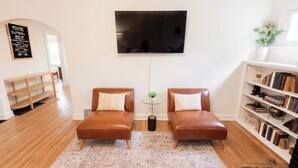 Una sala de estar con dos sofás de cuero, una mesa de centro, un televisor montado en la pared, una estantería y una escalera que lleva a otra habitación.