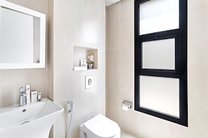 A modern bathroom with a white sink, toilet, and shower area. There is a window with black framing and a shelf with toiletries.