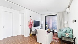 Una sala moderna con un sofá, una mesa de centro y un televisor. Hay un ventanal con cortinas, una planta y vistas de la ciudad.