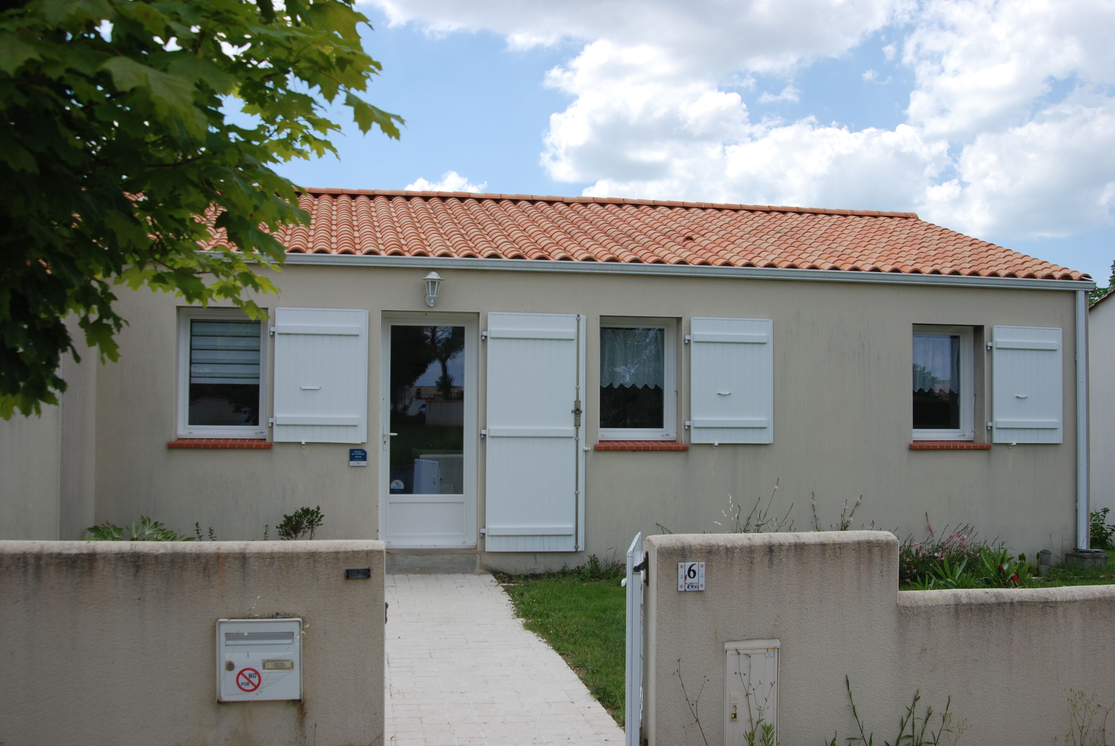Un bâtiment à deux étages, avec un toit recouvert de tuiles, des volets blancs et un petit jardin à l’avant.